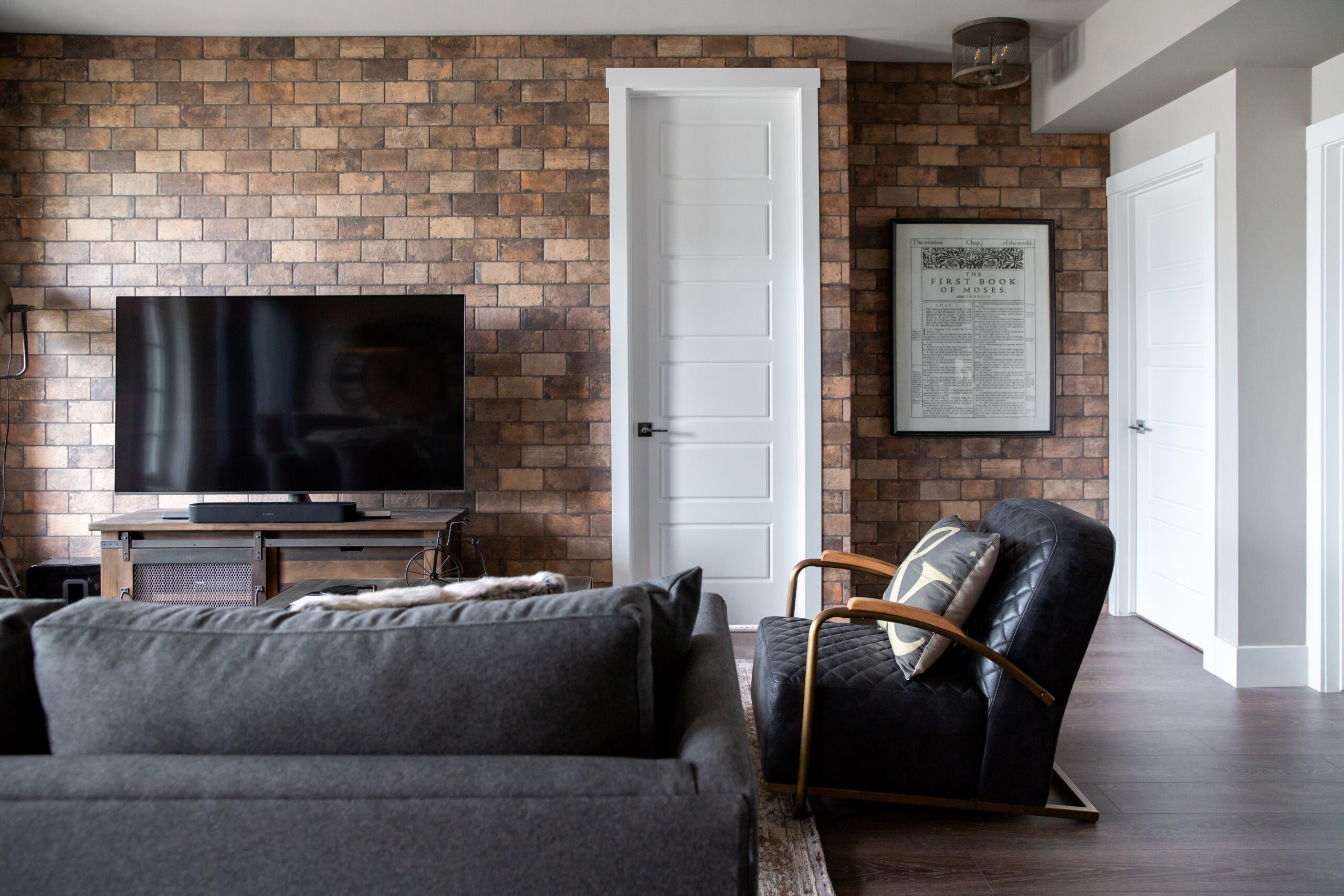 A living room in a condo decorated with masculine touches. It has a brick accent wall in the living room with a blue plush sofa, wood table and black leather chair.