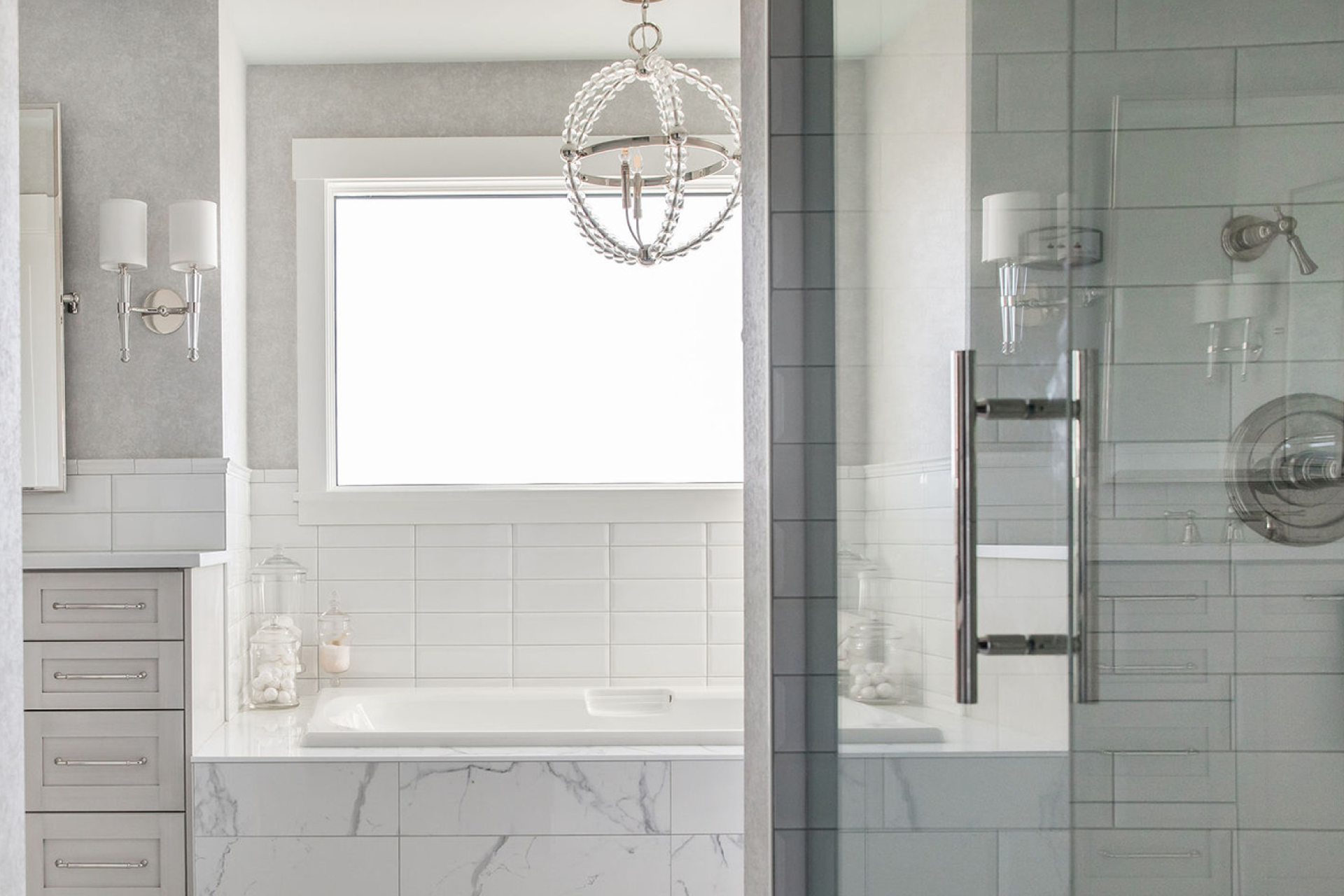 Unique design elements in white bathroom that include, marble, title, glass, chandelier lighting and stainless fixtures.