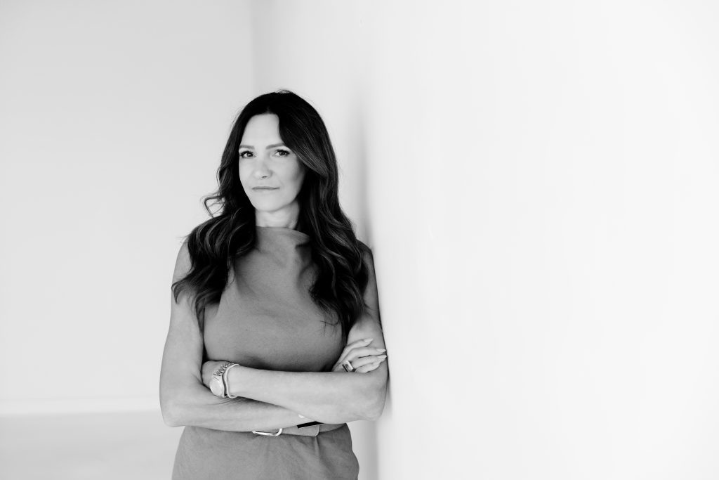 A black and white photo of a woman leaning against a wall with her arms crossed and facing the camera. She is in front of a light grey wall. She is a senior interior designer and the owner of Lush Interiors.