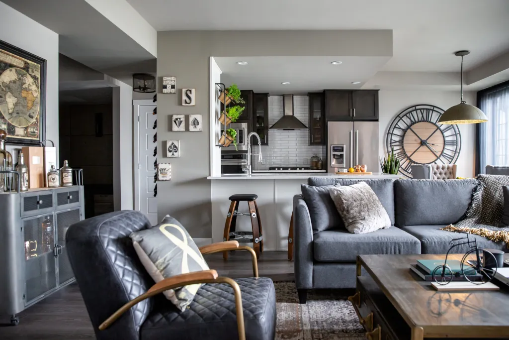 Elevated condo decor in a dining room and living room, in blue and brown tones with masculine touches like a giant wall clock with roman numerals, a metal wet bar, and whiskey barrel bar chairs.