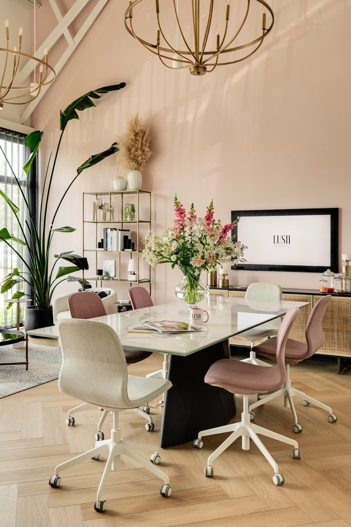 A meeting room with high ceilings, an ornate chandelier, pink walls, a video screen and a long table with a large bouquet of flowers.
