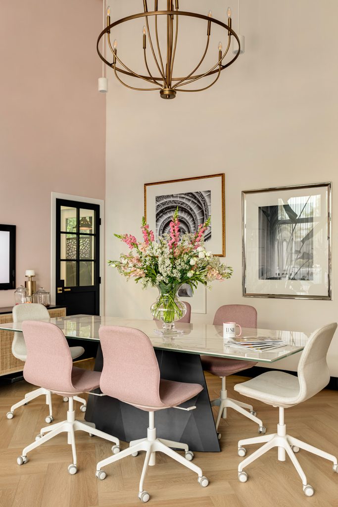 A meeting room with high ceilings, an ornate chandelier, pink accents, and a long table with a large bouquet of flowers.
